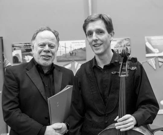 John Moore, conductor, standing next to and shaking hands with Graham Walker, soloist, who is holding his cello.