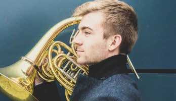 Ben Goldscheider facing left with his collar up and holding his French horn over his right shoulder.