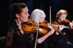 Zoë Beyers seated and playing her violin. There are two other violin players beyond her.